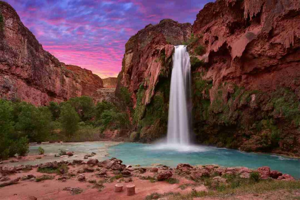 a waterfall flows from red rocks with a purple sunset