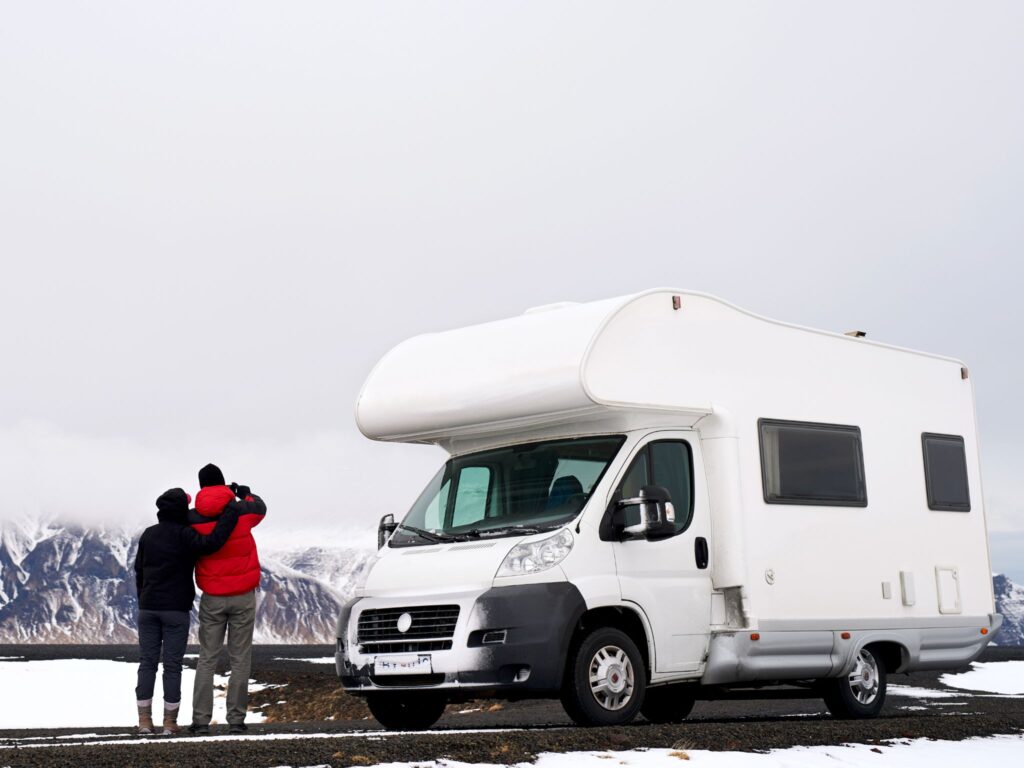 A couple stands beside their rv while looking at snow-capped mountains